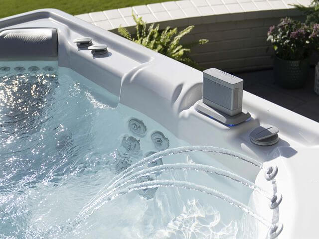 Close-up of a luxurious outdoor hot tub with arcing water jets, built-in speakers, and ergonomic seating, set on a patio with greenery and flowers.